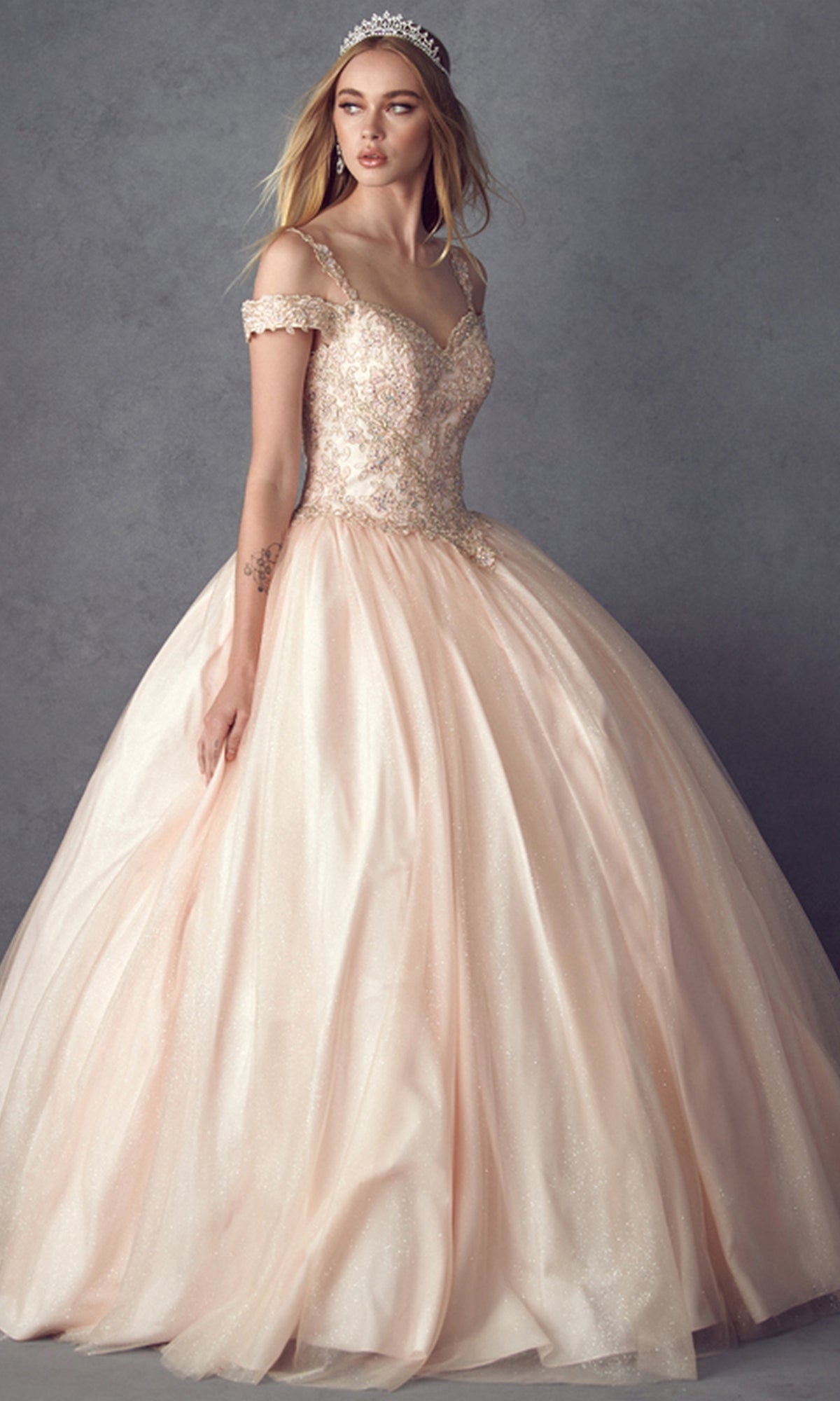 Wearing the Juliet 1430 Cold-Shoulder Glitter Quinceanera Dress and a sparkling tiara, a woman stands against a plain gray background.